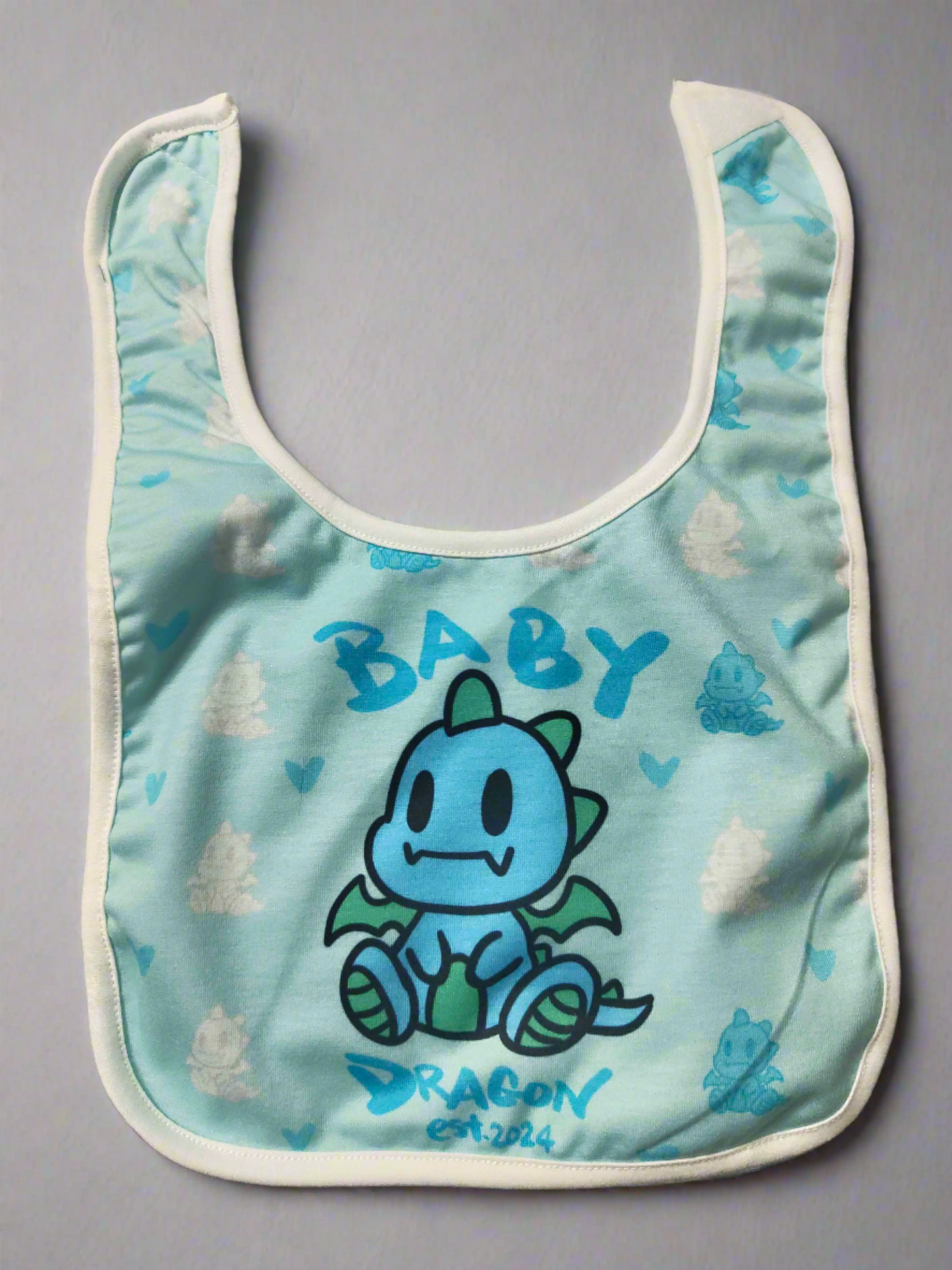 A light blue baby bib with a white border. It features a cartoon blue dragon in the center with the word "BABY" written above it and "DRAGON est. 2024" written below it.
