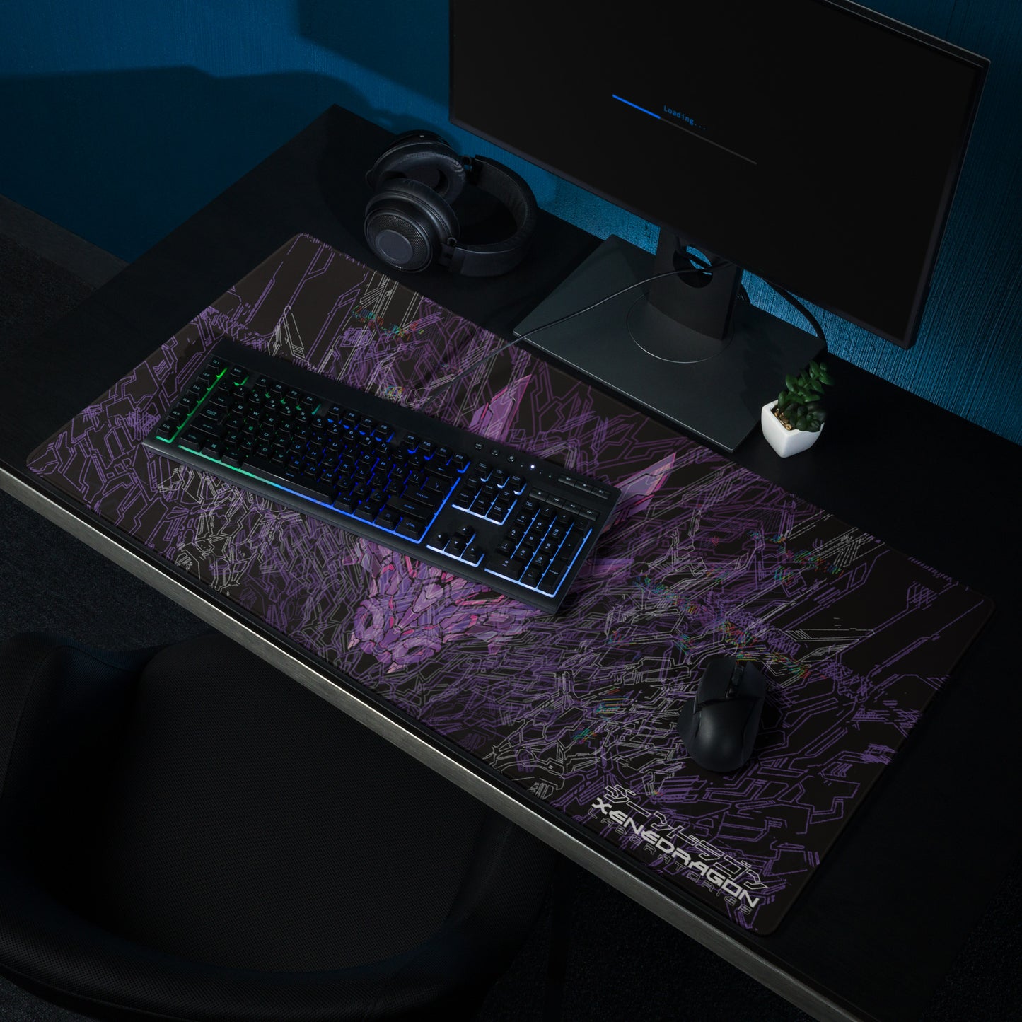 A black mousepad with a large, detailed illustration of a dragon's head in shades of purple. and wireframe design. The word "XENEDRAGON LABORATORIES" is written in small font at the bottom of the mousepad. The mousepad is shown on a desk with a keyboard, mouse, headphones, and a small plant.