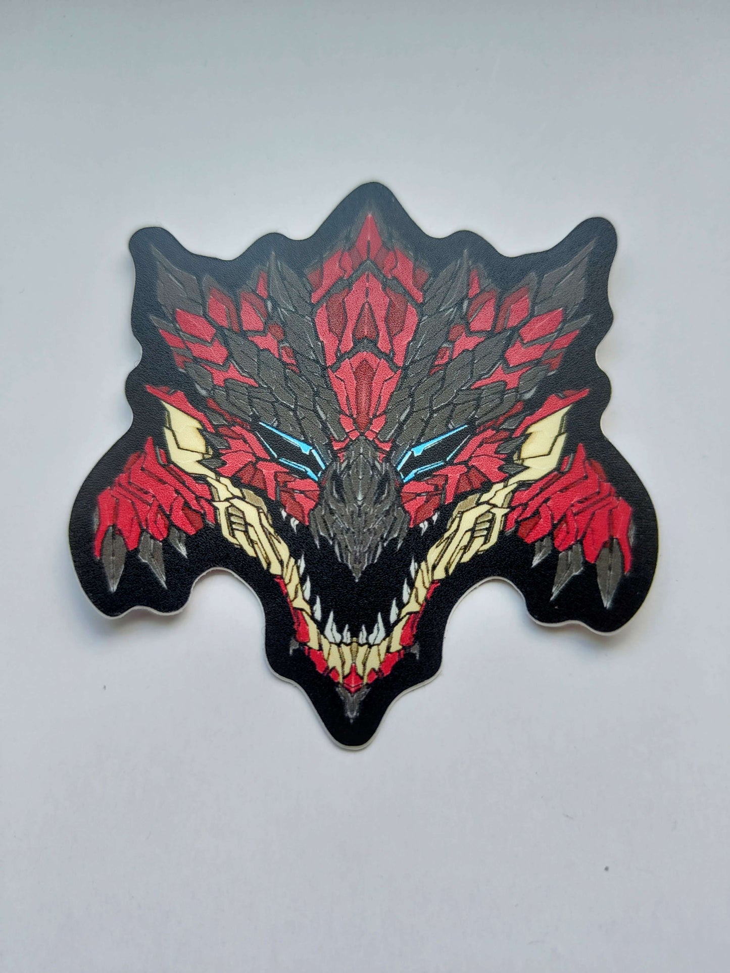 A sticker with a detailed design of a stylized monster's head. The design features vibrant red themes with light blue eyes. Also dark themes like black and grey for the scales and horns. Designed by XeneDragon