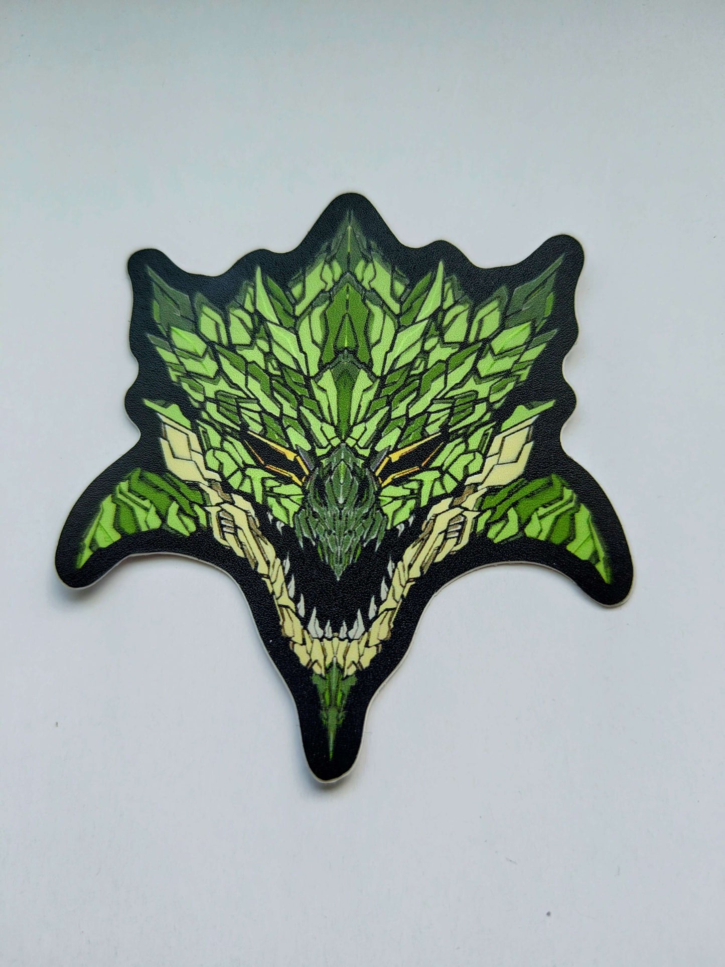 A sticker with a detailed design of a stylized monster's head. The design features green themes with yellow blue eyes. Also darker greens for the scales and horns. Designed by XeneDragon.