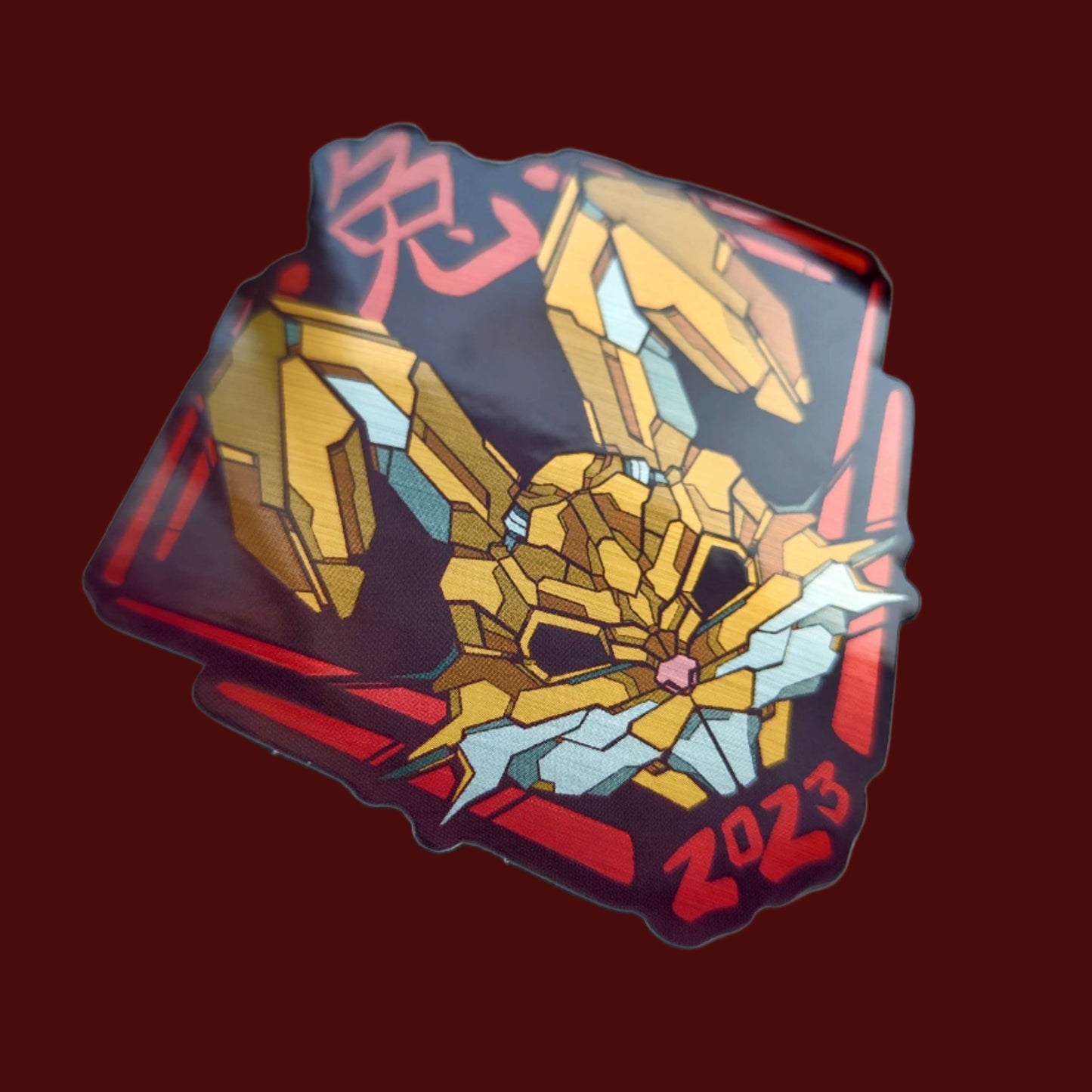 A special metallic effect sticker with a vibrant geometric design celebrating the Year of the Hare. The sticker features a stylized image of a hare's head in gold, with the Chinese character for "hare" and the year "2023" printed below in red. Designed by XeneDragon