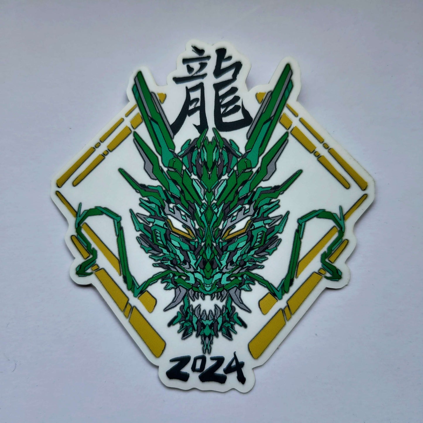 A special clear effect sticker with a vibrant geometric design celebrating the Year of the Dragon. The sticker features a stylized image of a dragon's head in green, with the Chinese character for "dragon" and the year "2024" printed below in yellow. Designed by XeneDragon