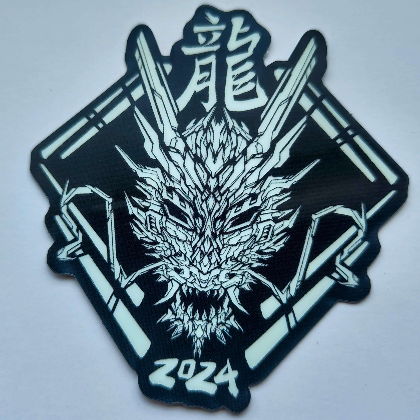 A special glow in the dark effect sticker with a geometric design celebrating the Year of the Dragon. The sticker features a stylized image of a dragon's head, with the Chinese character for "dragon" and the year "2024" printed below. Designed by XeneDragon