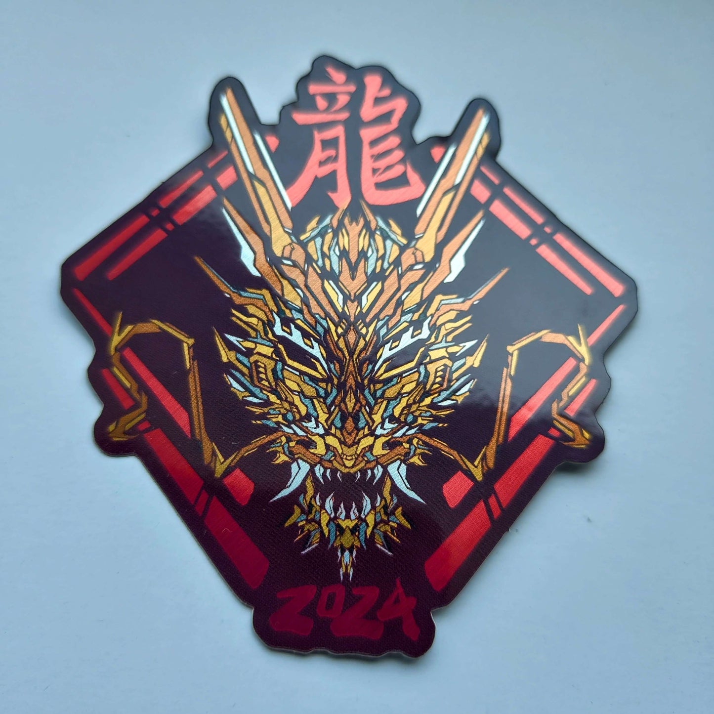 A special metallic effect sticker with a vibrant geometric design celebrating the Year of the Dragon. The sticker features a stylized image of a dragon's head in gold, with the Chinese character for "dragon" and the year "2024" printed below in red. Designed by XeneDragon