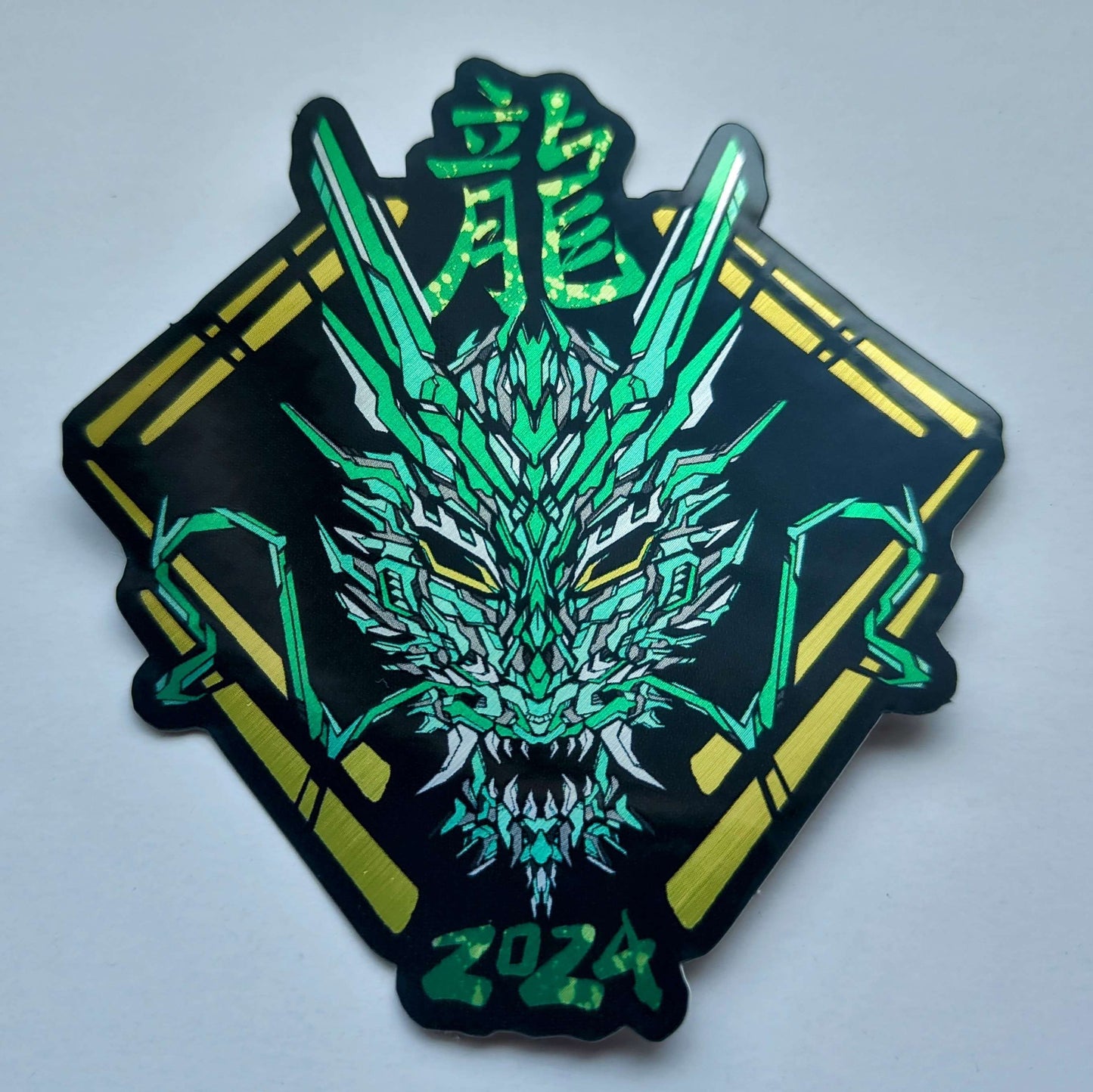 A special metallic effect sticker with a vibrant geometric design celebrating the Year of the Dragon. The sticker features a stylized image of a dragon's head in green, with the Chinese character for "dragon" and the year "2024" printed below in yellow. Designed by XeneDragon