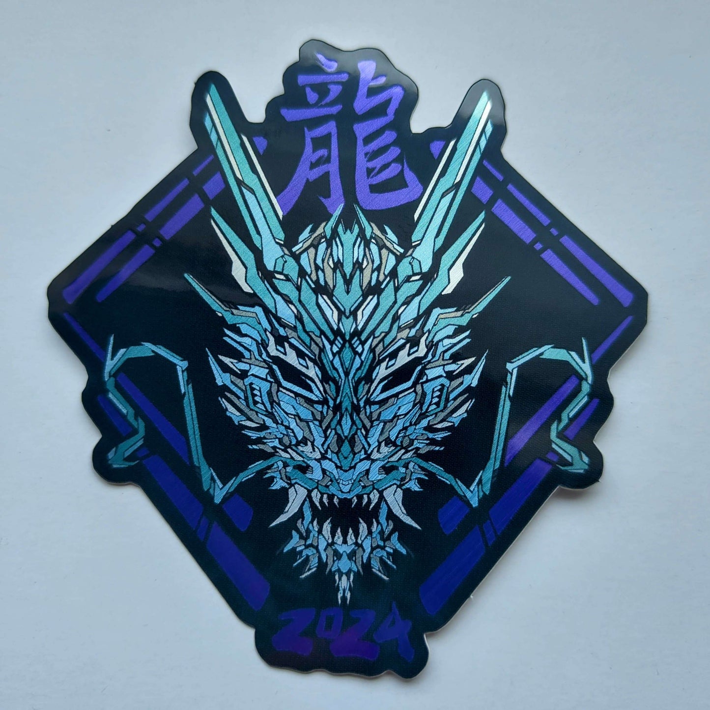 A special metallic effect sticker with a vibrant geometric design celebrating the Year of the Dragon. The sticker features a stylized image of a dragon's head in blue, with the Chinese character for "dragon" and the year "2024" printed below in purple. Designed by XeneDragon