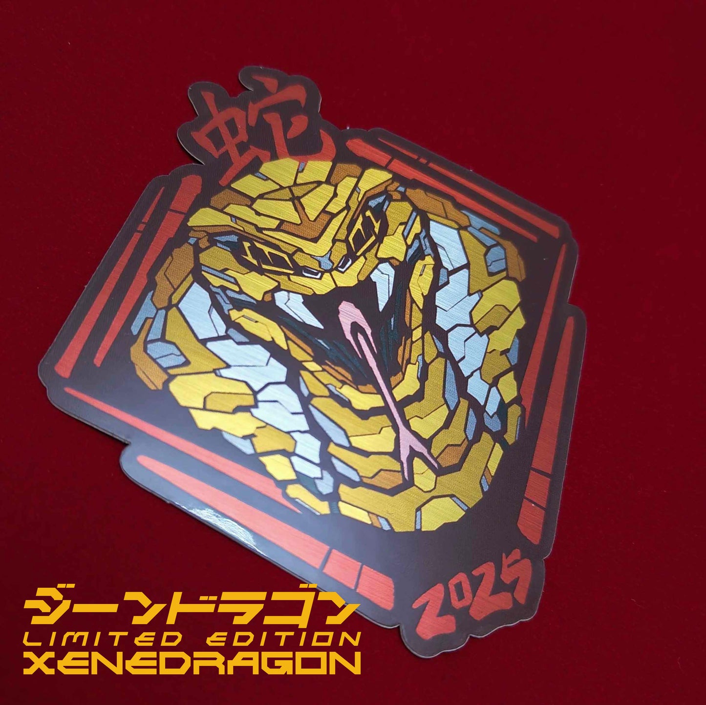 A special metallic effect sticker with a vibrant geometric design celebrating the Year of the Snake. The sticker features a stylized image of a cobras head in gold, with the Chinese character for "snake" and the year "2025" printed below in red. Designed by XeneDragon
