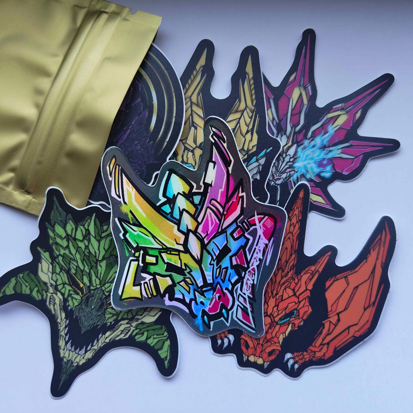 a collection of stickers featuring various dragon-like creatures. The stickers are colorful and have a detailed, almost graffiti-like style. They are arranged on a surface with a gold foil bag in the background.