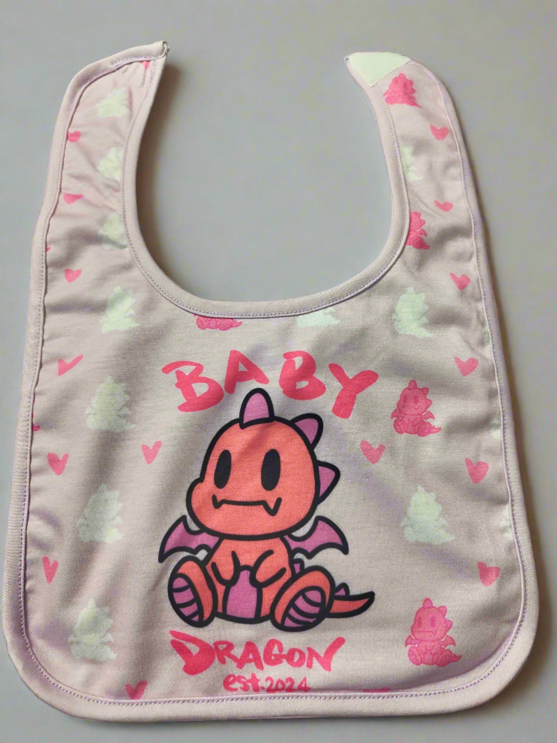 A pink baby bib with a pink border. It features a cartoon pink dragon in the center with the word "BABY" written above it and "DRAGON est. 2024" written below it.