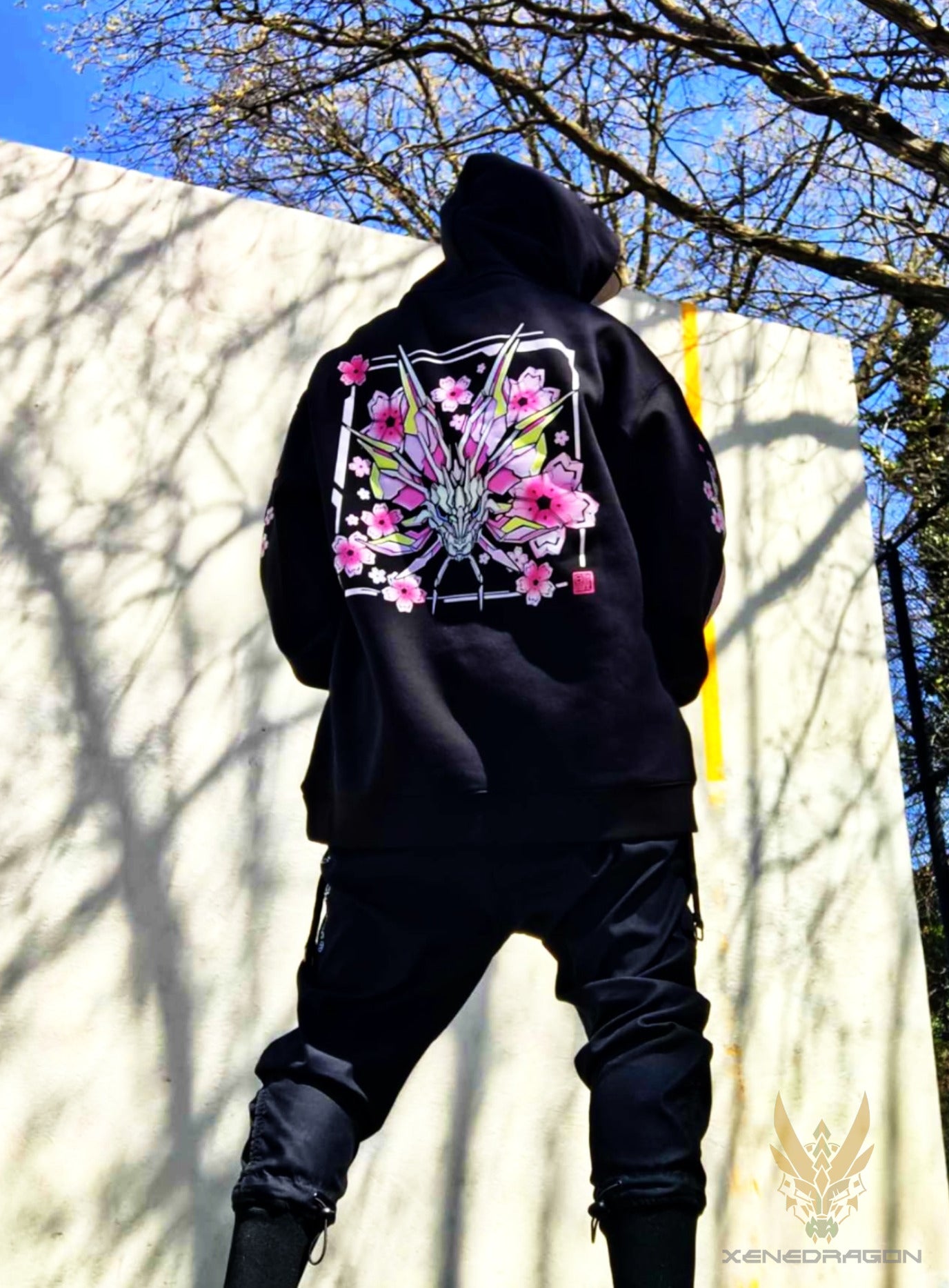 A picture of a model wearing a hoodie in the park with blue sky and trees in the background. The model is facing away to showcase the back of the design
