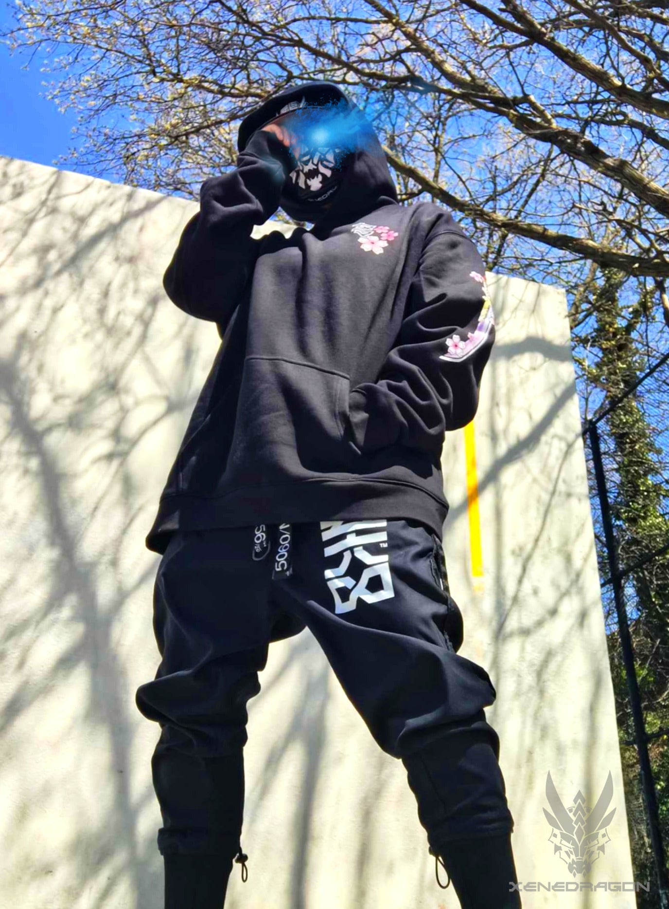 A picture of a model wearing a hoodie in the park with blue sky and trees in the background. The picture has an edited flaming blue eye on the model 
