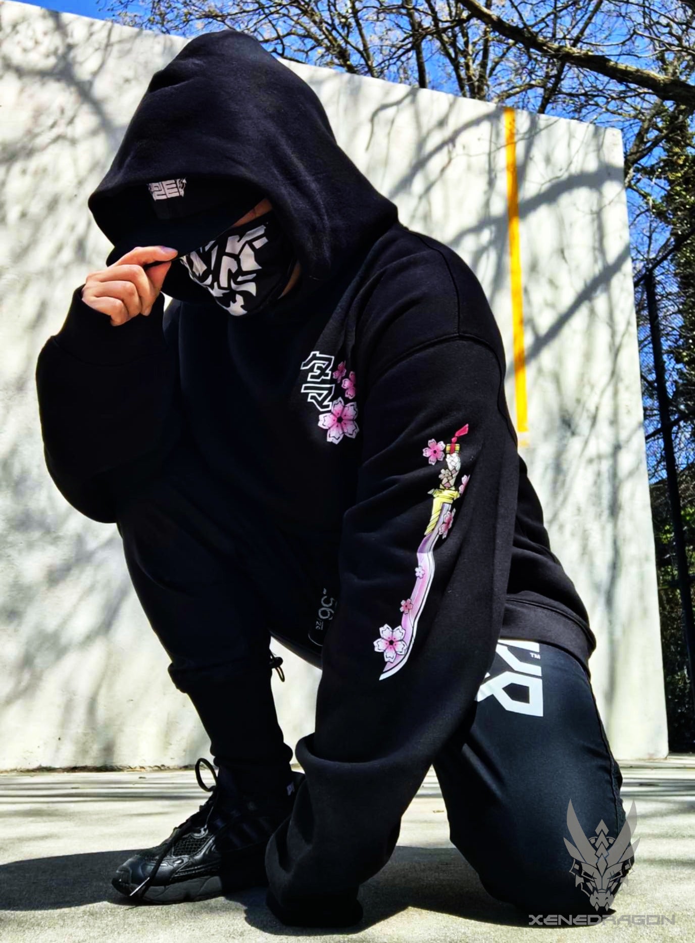 A picture of a model wearing a hoodie in the park with blue sky and trees in the background. The model is crouching down to show case the design on the sleeve