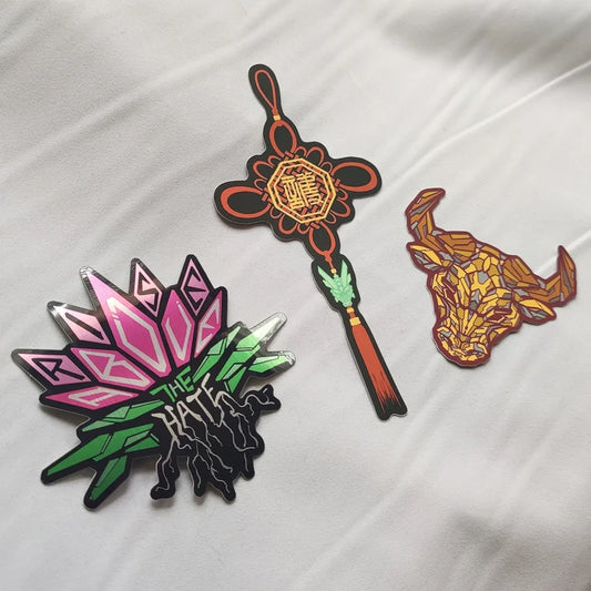 Three colorful stickers displayed on a white surface. One features a pink lotus flower design, another has a Chinese knot symbol, and the third depicts a golden bull's head.